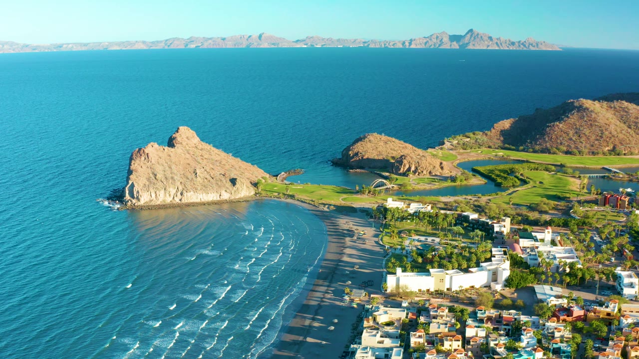 A wide aerial view of Loreto coastline shows small surf lines approaching the beach, the rocky outcrop left of center, green shorefront grounds, clustered buildings at the lower right, and no visible people.