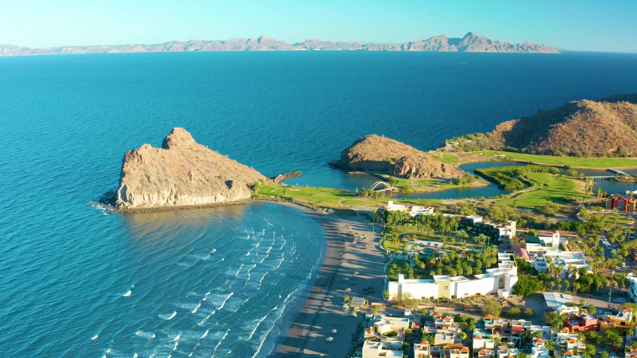 Coastal aerial view with rocky outcrop, beach surf, greenery, and buildings.