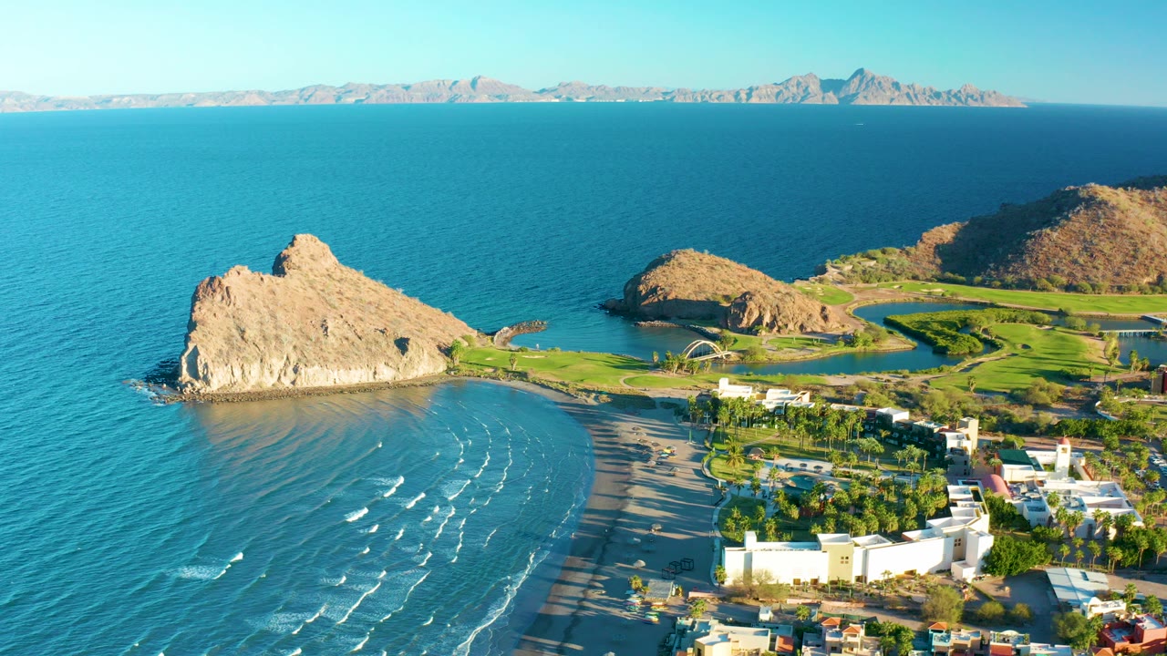 Opening aerial frame over Loreto, Baja California Sur, with blue ocean at left, a curved beach below, tan rock formations near shore, white buildings at right, distant mountains, and no visible people.