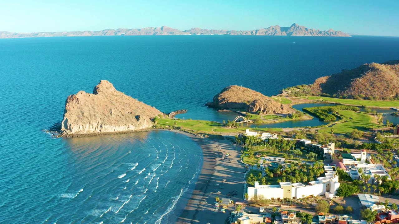 High coastal view with waves crossing a sandy beach beside rocks and low buildings.