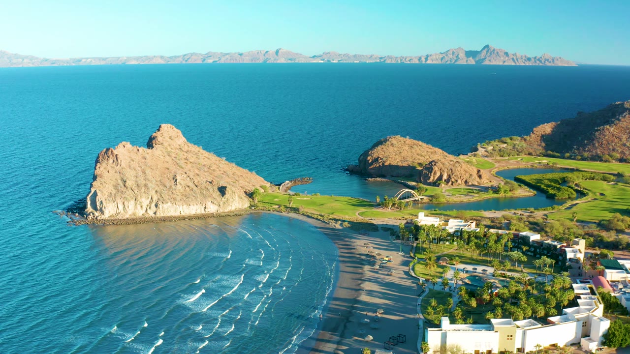 Aerial view in Loreto, Baja California Sur with the rocky point centered left, wave lines crossing the shallow bay, green grounds to the right, and no visible people.