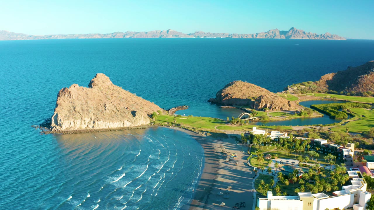 Wide aerial frame of the Loreto coast with a tan rock outcrop, bright blue ocean, a sandy beach, green coastal grounds, distant mountain ridges, and no visible people.