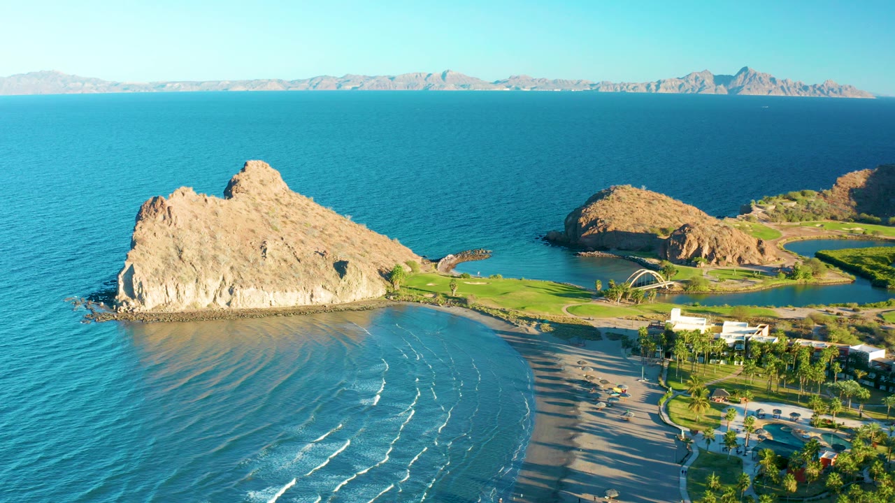 Aerial view of rocky outcrop, beach, blue ocean, and distant mountains.