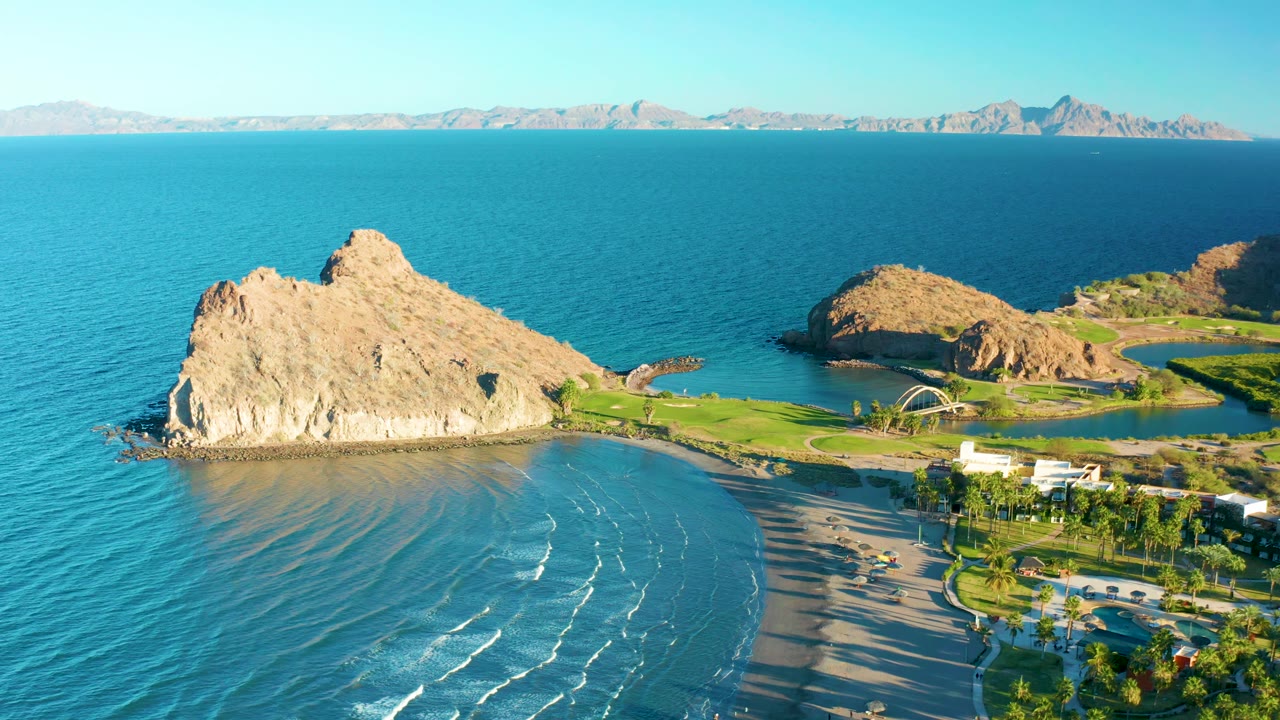 Aerial view in Loreto with the large rock slightly left of center, white wave lines bending toward the sandy shore, green grounds to the right, and no visible people.