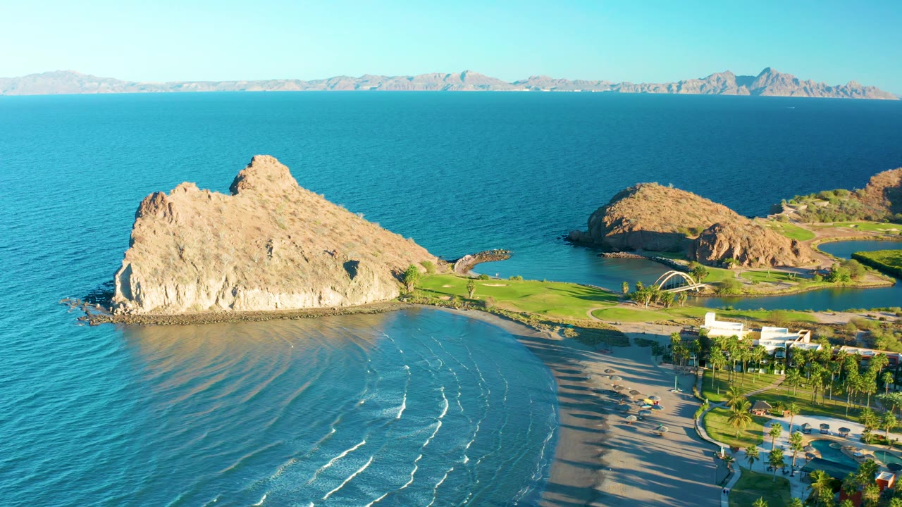 Wide aerial frame over Loreto coastline with more open water visible around the rocky outcrop, a sandy beach curving below, distant mountains, and no visible people.