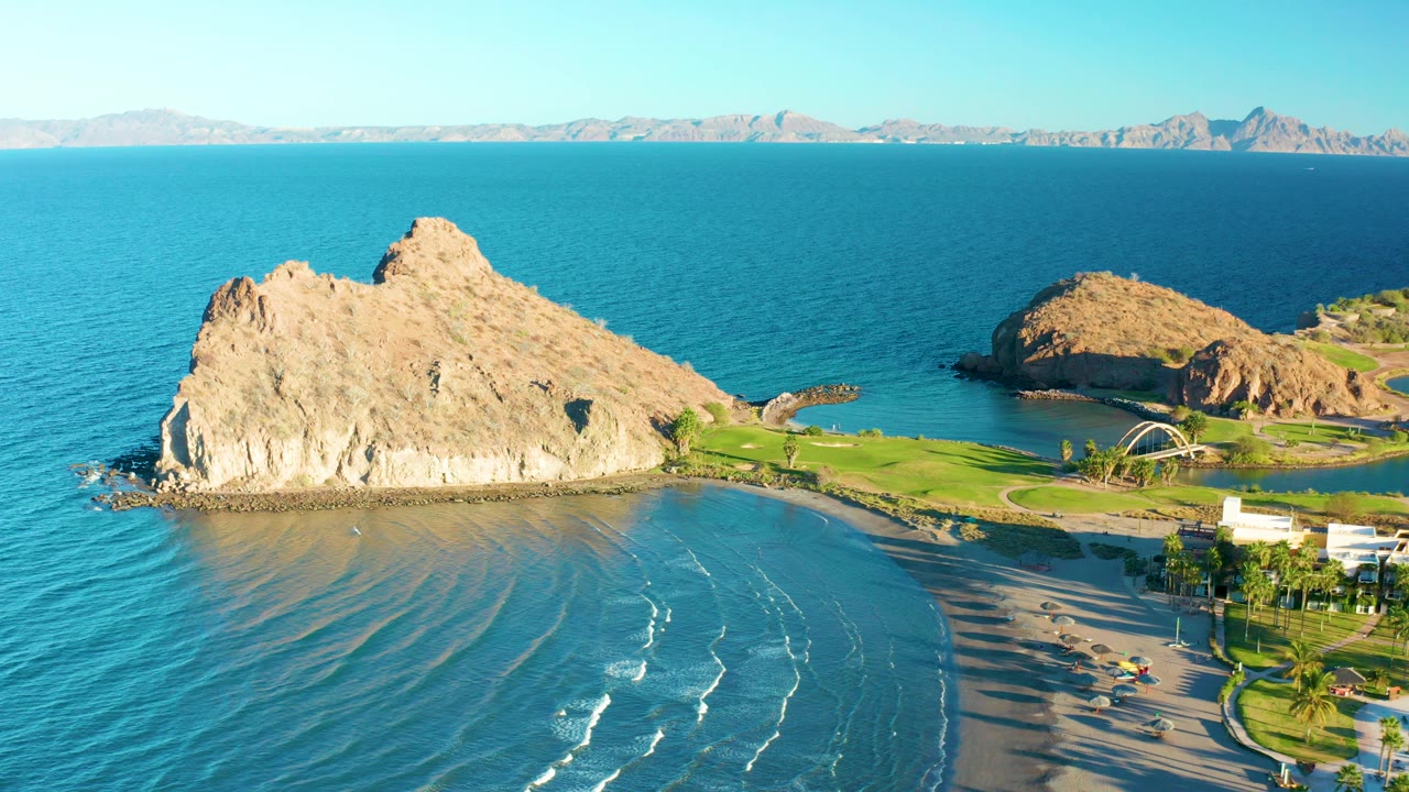 Aerial view in Loreto, Baja California Sur, showing a tan rocky outcrop, curved sandy beach, gentle surf lines, green grass, distant mountains, and tiny people near the lower beach.