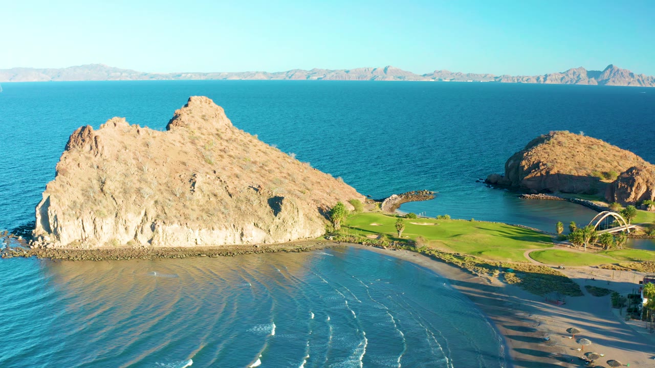 Aerial frame over Loreto showing the main rock formation slightly farther right, wave lines on the shallow beach water, a green shoreline strip, distant mountains, and no visible people.