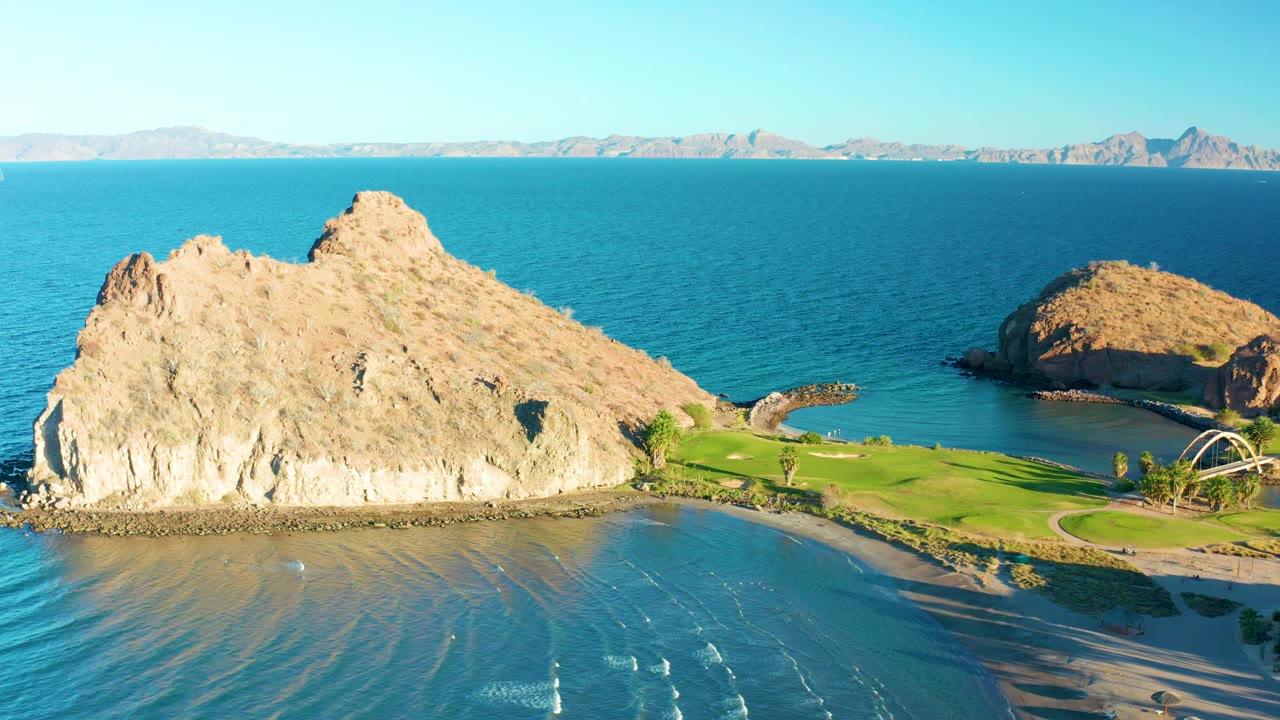 Mid-clip aerial frame over Loreto, Baja California Sur, with the large rock occupying the left half, pale surf lines in shallow water below, a green strip and path at right, and no visible people.