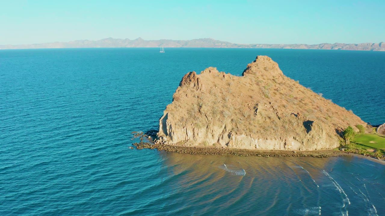 Aerial view of a tan rocky island off Loreto, Baja California Sur, surrounded by blue ocean with distant mountains, a small white object near the left waterline, and no visible people.