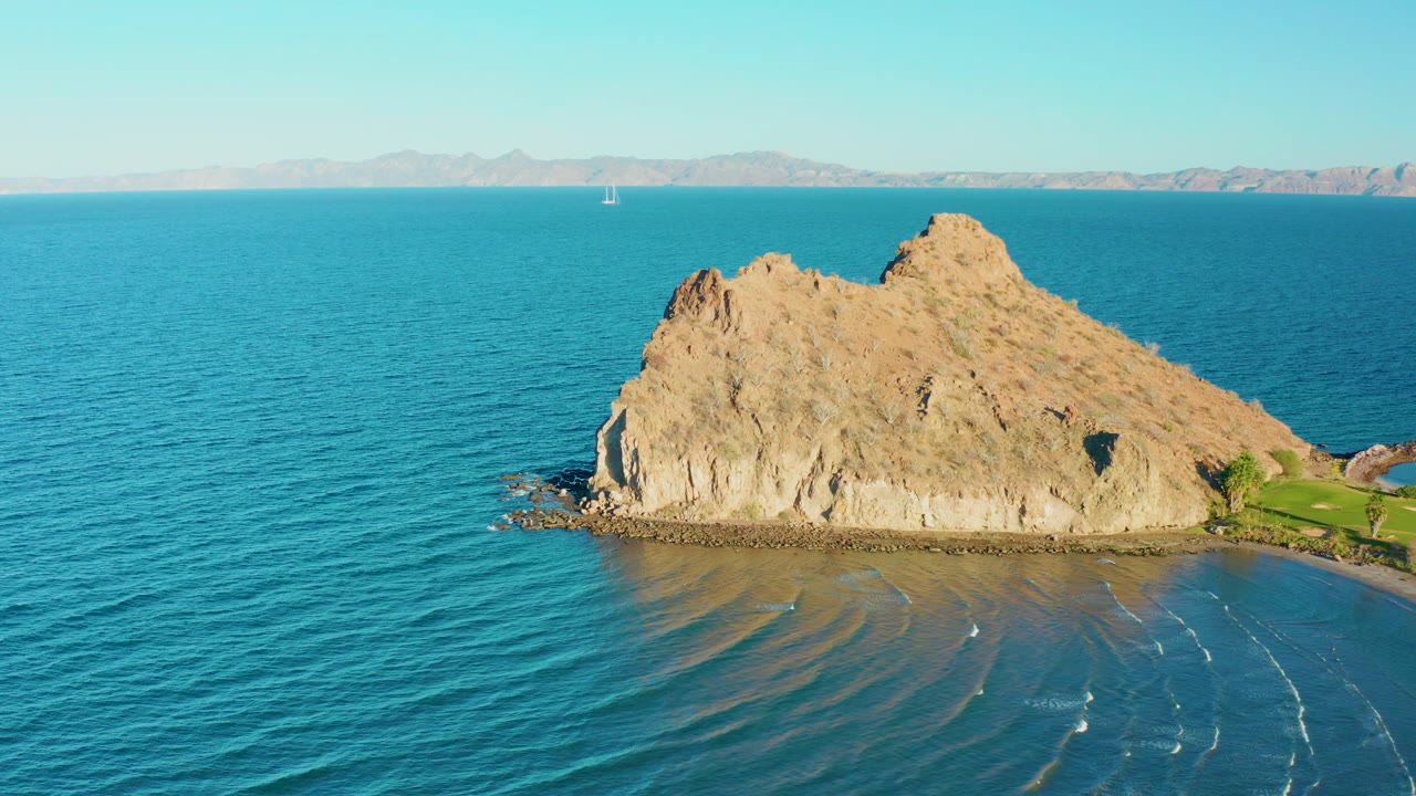 Aerial frame in Loreto, Baja California Sur with the rocky island centered, calm blue sea spreading across the foreground, a pale mountain horizon, and no visible people.