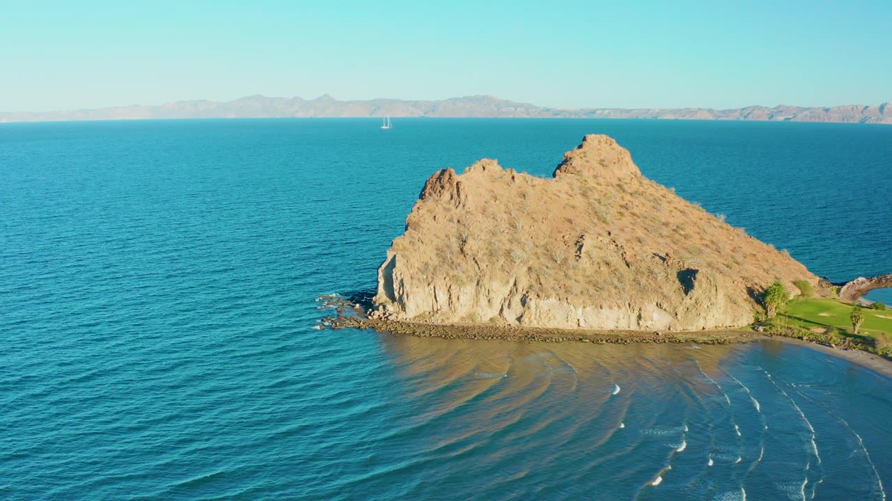 Loreto, Baja California Sur aerial still showing the island slightly right of center, blue ocean in front, thin white wave traces near shore, distant mountains, and no visible people.