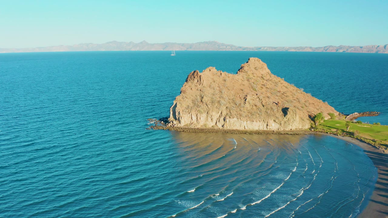 Aerial frame in Loreto, Baja California Sur showing the rock slightly more centered, fine surf lines on the curved beach, distant mountains, and no visible people.