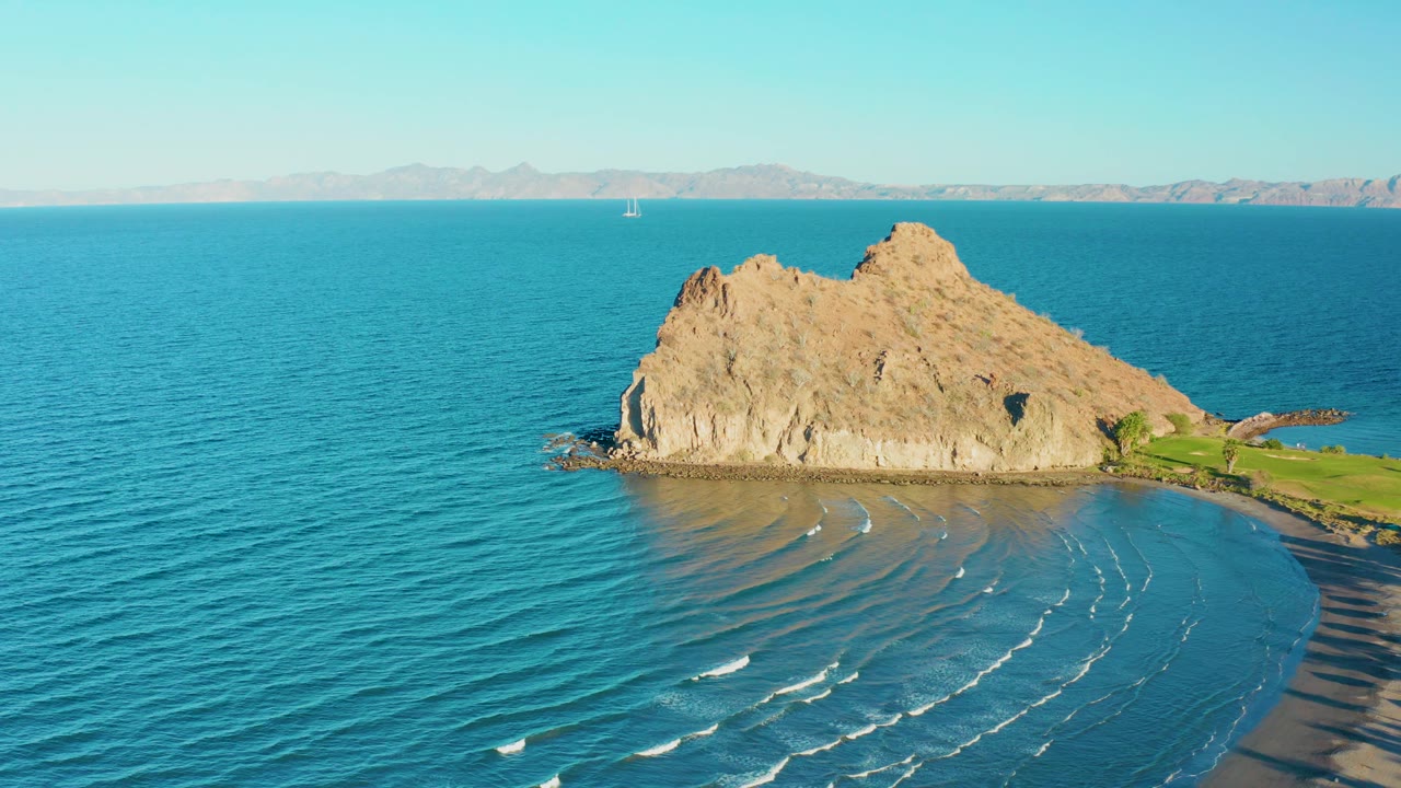Aerial frame in Loreto, Baja California Sur with more open water visible at left, the rocky outcrop near center, a sandy shoreline at lower right, and no visible people.