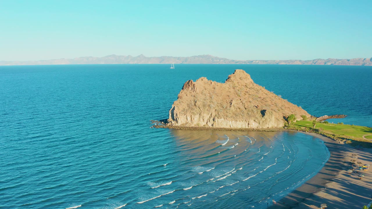 Opening aerial frame over Loreto, Baja California Sur showing a tan rocky outcrop, turquoise water, curved sandy beach, distant sailboat, and no visible people.