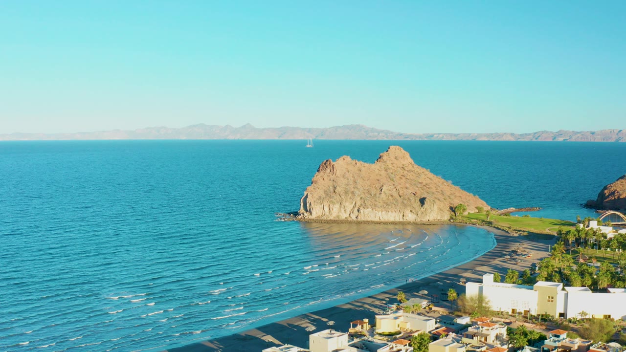 Opening aerial frame over Loreto, Baja California Sur, showing turquoise water, a tan rocky outcrop, curved beach, distant coast, and town rooftops with no visible people.