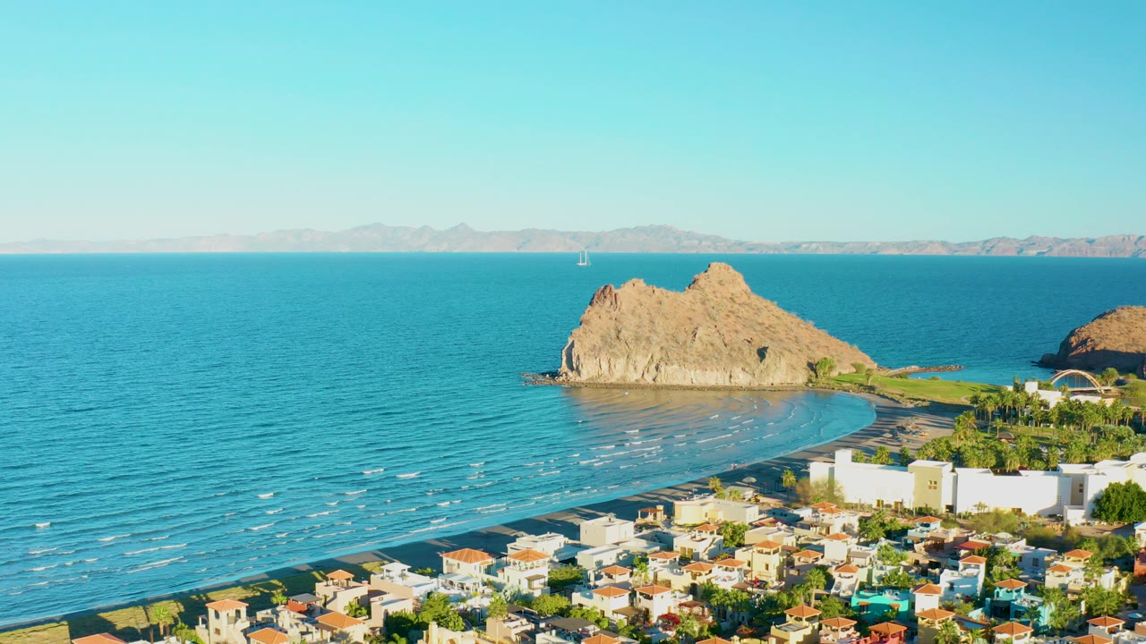 Opening frame over Loreto, Baja California Sur shows town rooftops below a curved beach, calm blue water, rocky outcrops, distant mountains, clear sky, and no visible people.