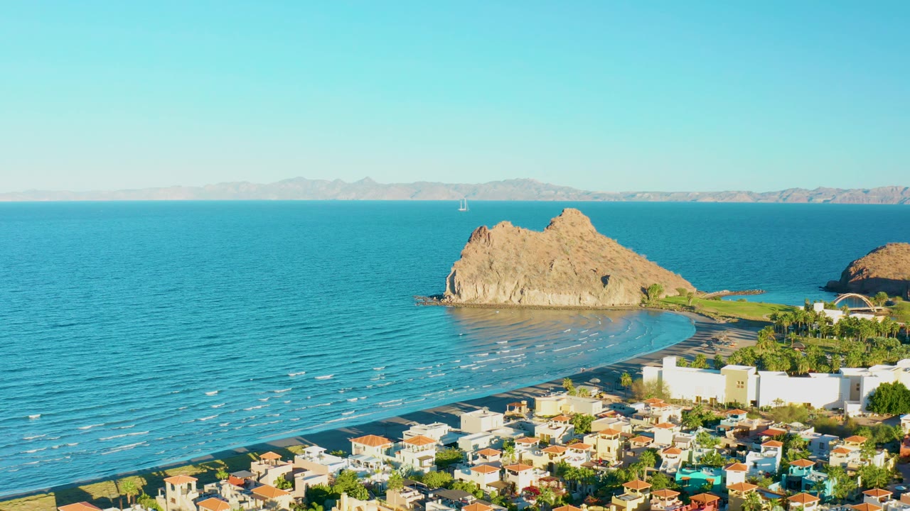 At 0:01, Loreto town buildings fill the lower frame while the beach, calm sea, rocky outcrop, faint distant mountains, and clear sky remain visible with no visible people.