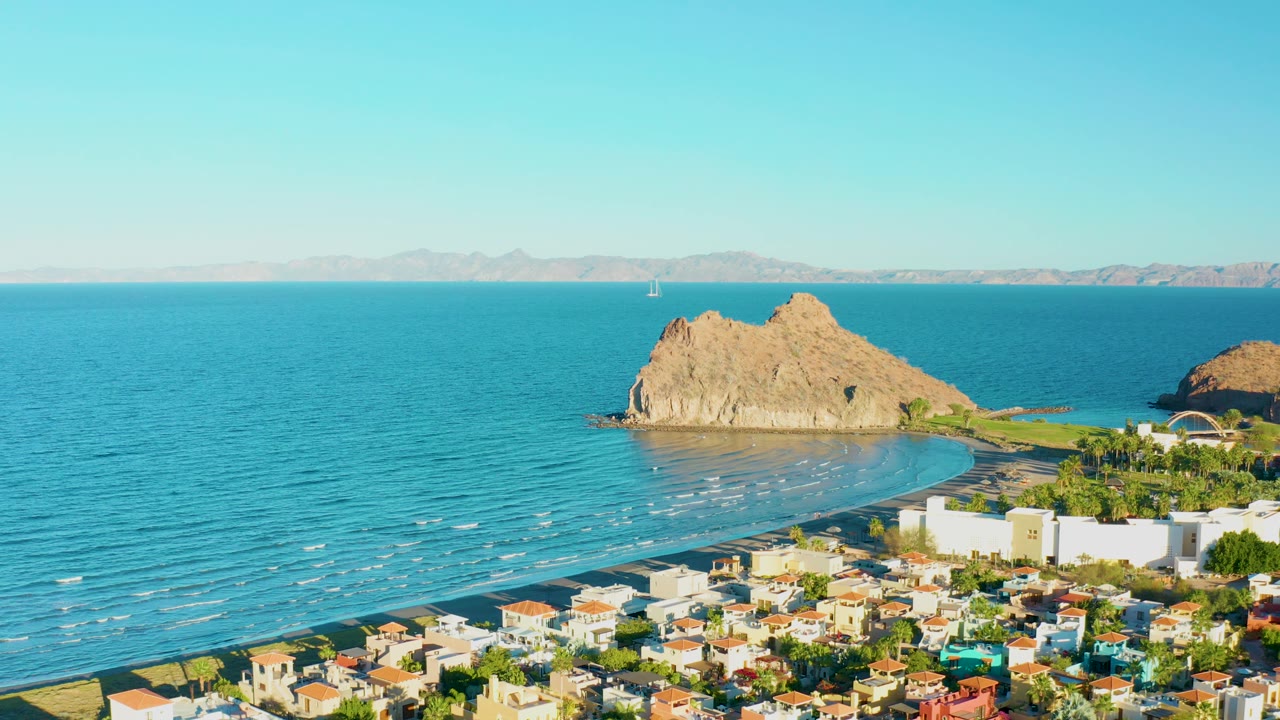 At 0:02, the rocky outcrop sits near the center of the Loreto coastline, with turquoise water, a pale beach curve, compact rooftops, distant mountains, clear sky, and no visible people.