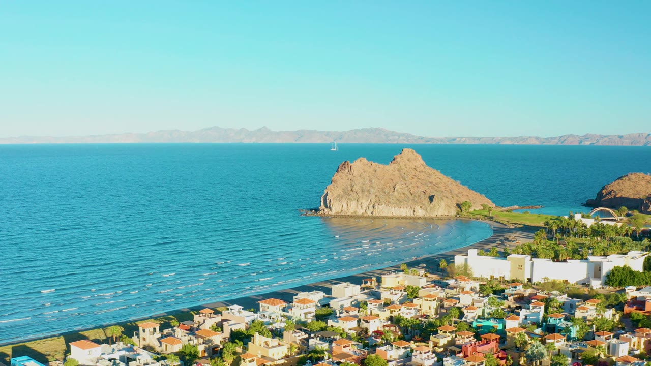 Elevated view of beach town, calm ocean, rocky outcrops, and distant mountains.