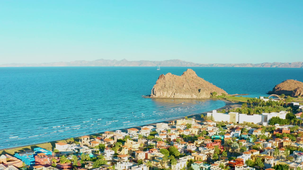 Aerial frame over Loreto, Baja California Sur, showing colorful rooftops below a blue sea with tan rock formations near shore and no visible people.