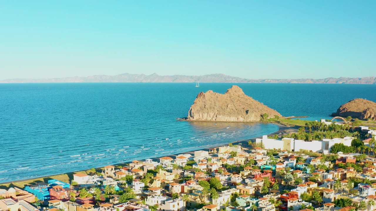 Loreto coastal rooftops fill the foreground while calm blue sea, narrow white wave lines, tan rocks, and a pale horizon sit beyond, with no visible people.