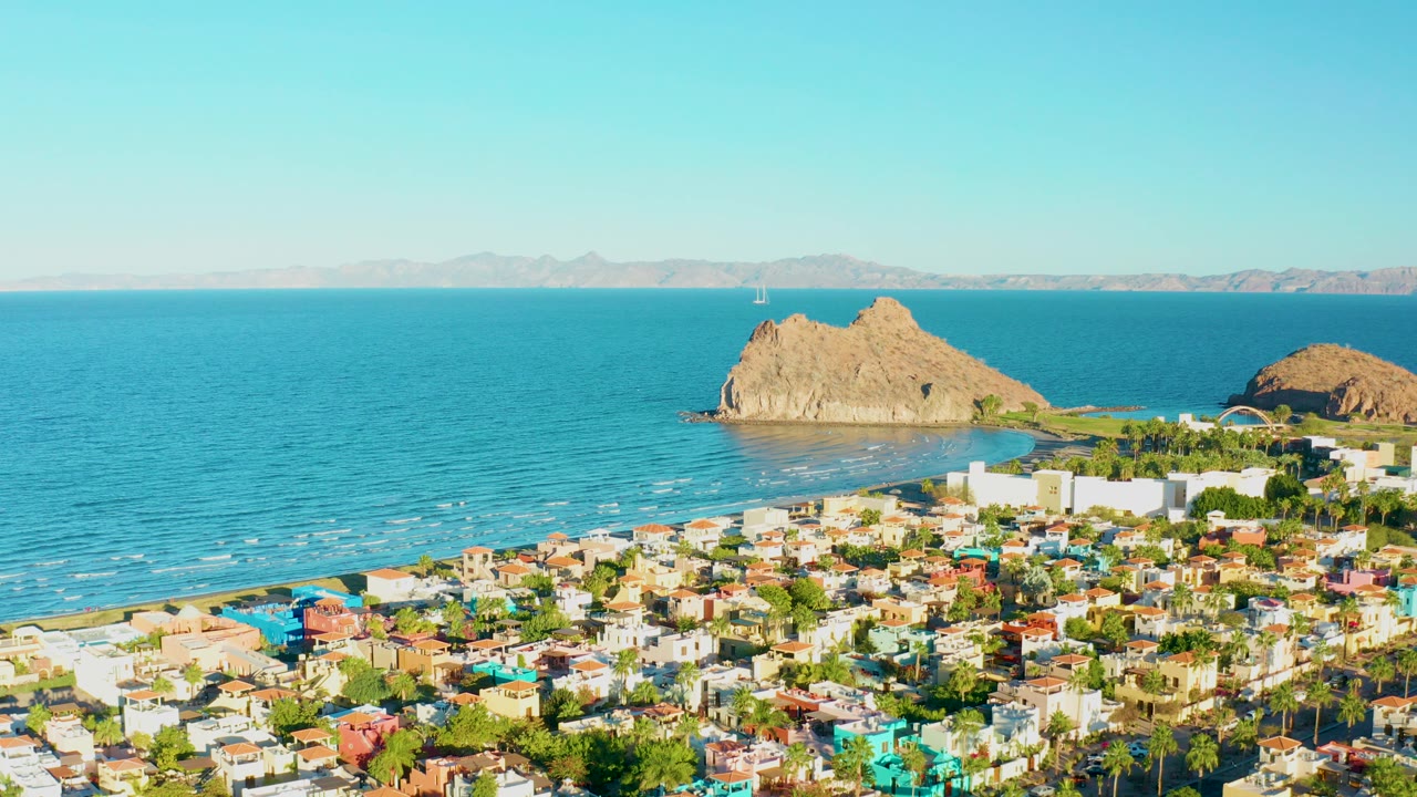 Opening aerial view of Loreto, Baja California Sur with colorful low-rise buildings below, blue ocean beyond the beach, tan rock formations offshore, clear sky, and no visible people.