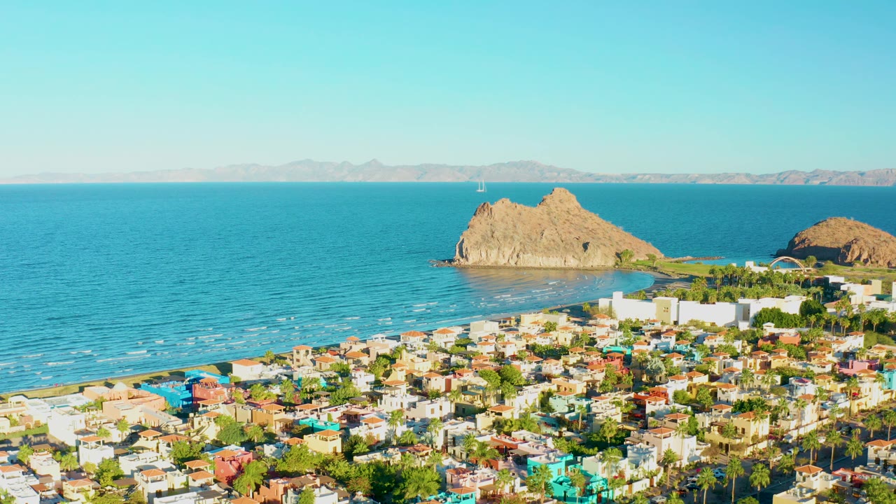 At 0:01, the Loreto aerial holds on dense colorful rooftops, palm-lined blocks, the pale beach edge, rippled blue water, distant land, and no visible people.