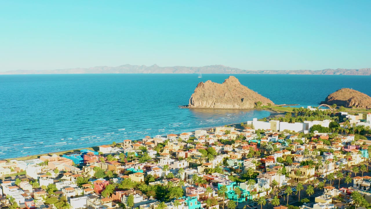 At 0:02, the frame keeps the Loreto town in the lower foreground while turquoise water, faint surf lines, two tan offshore rock forms, distant land, and clear sky fill the upper view, with no visible people.