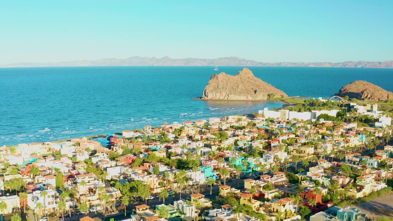 Opening frame over Loreto, Baja California Sur, with colorful low-rise buildings in front, blue ocean and rocky islands beyond, distant mountains on the horizon, and no visible people.