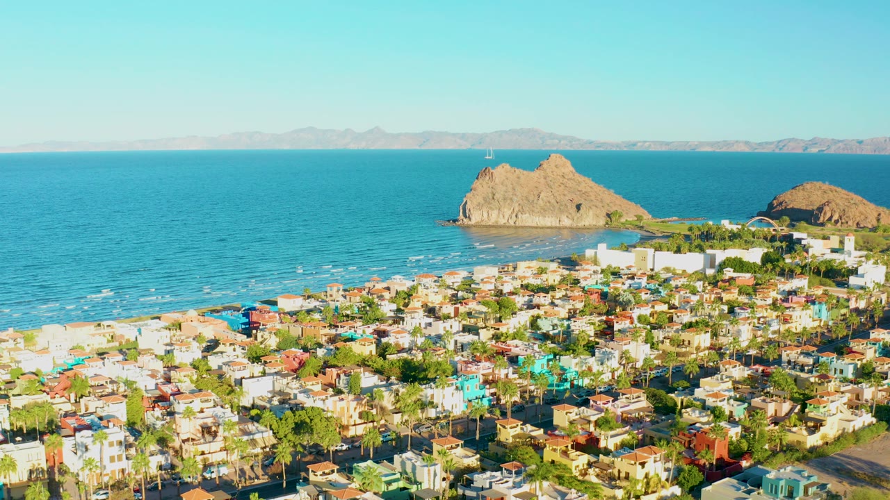 High aerial view across town rooftops toward blue water and two rocky landforms.