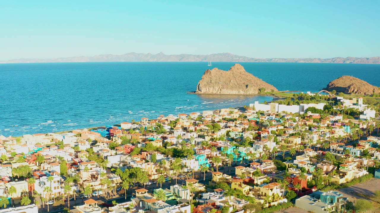 At 0:02, Loreto's coastal neighborhood fills the foreground, the sandy beach and calm blue ocean stretch behind it, and rocky islands sit under a clear sky with no visible people.