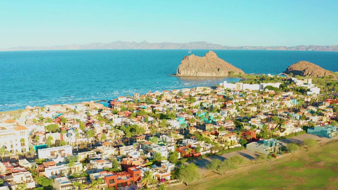 Opening aerial frame over Loreto, Baja California Sur, showing colorful low buildings, green grass, a sandy beach, blue ocean, two rocky outcrops, distant mountains, and no visible people.