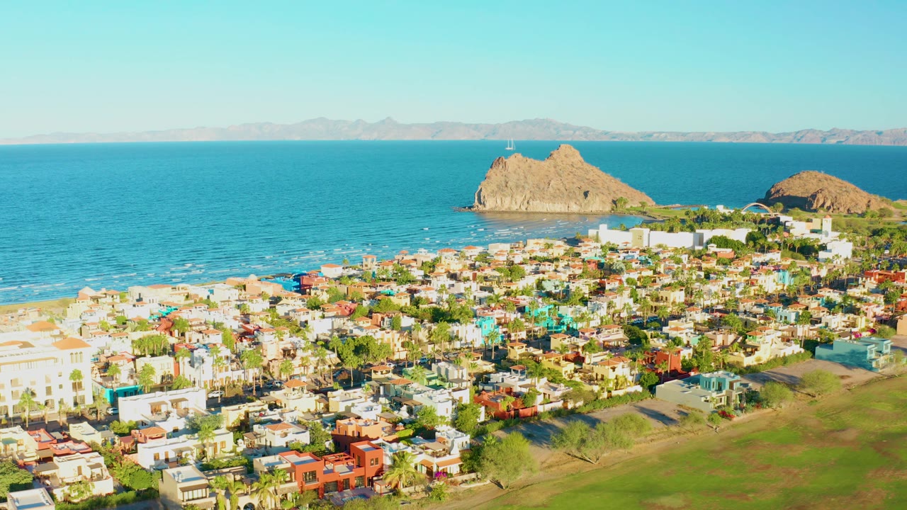 Aerial view of Loreto, Baja California Sur, showing bright low buildings, green foreground grass, beach, blue ocean, rocky outcrops, a distant mountain horizon, and no visible people.