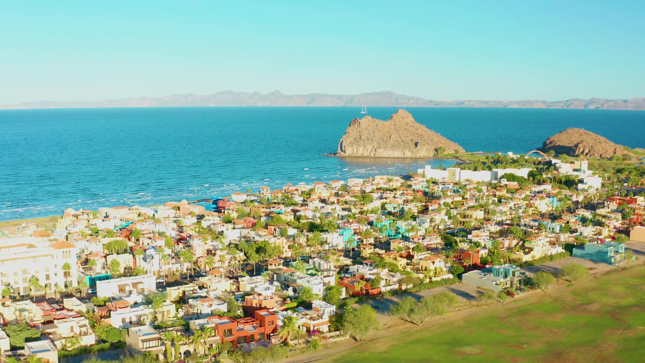 Aerial coastal view with town rooftops, green grass, beach, and rocky outcrops offshore.