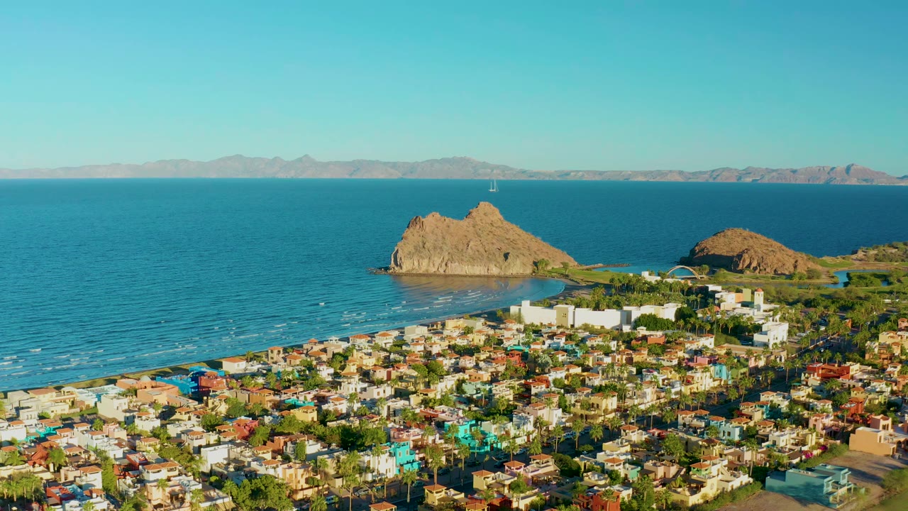 Opening aerial frame over Loreto, Baja California Sur, with colorful houses below, blue ocean beyond, rocky outcrops near shore, distant mountains, clear sky, and no visible people.