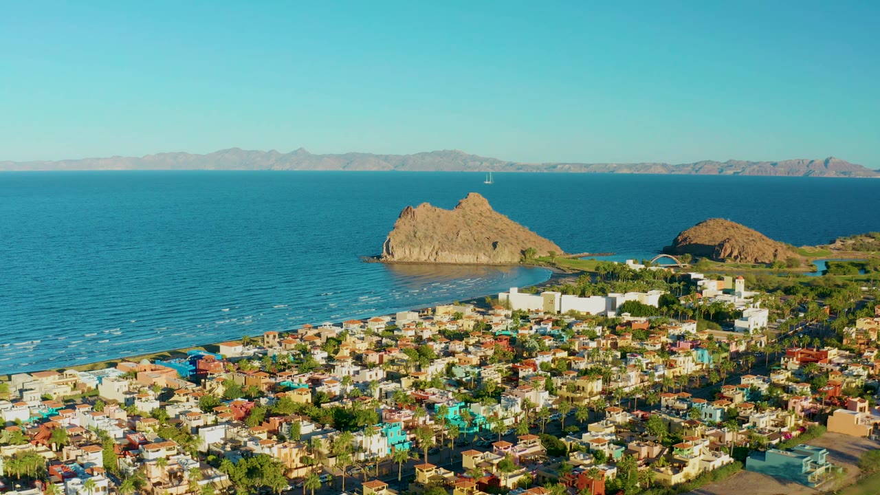 Aerial view over Loreto with a dense coastal neighborhood in the lower frame, turquoise-blue water across the middle, tan rocky islands near the shore, far mountains, and no visible people.