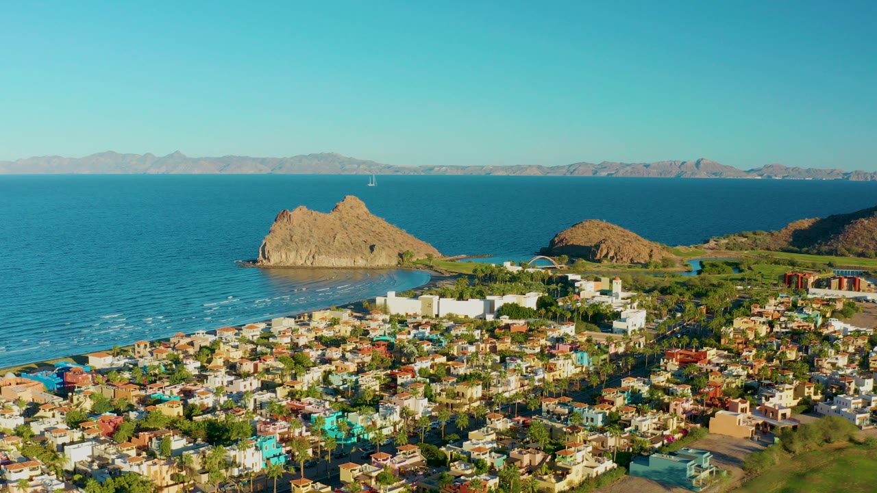 Aerial view over Loreto, Baja California Sur, with colorful low-rise buildings, palms, blue ocean, a tan rocky outcrop, distant mountains, and no visible people.