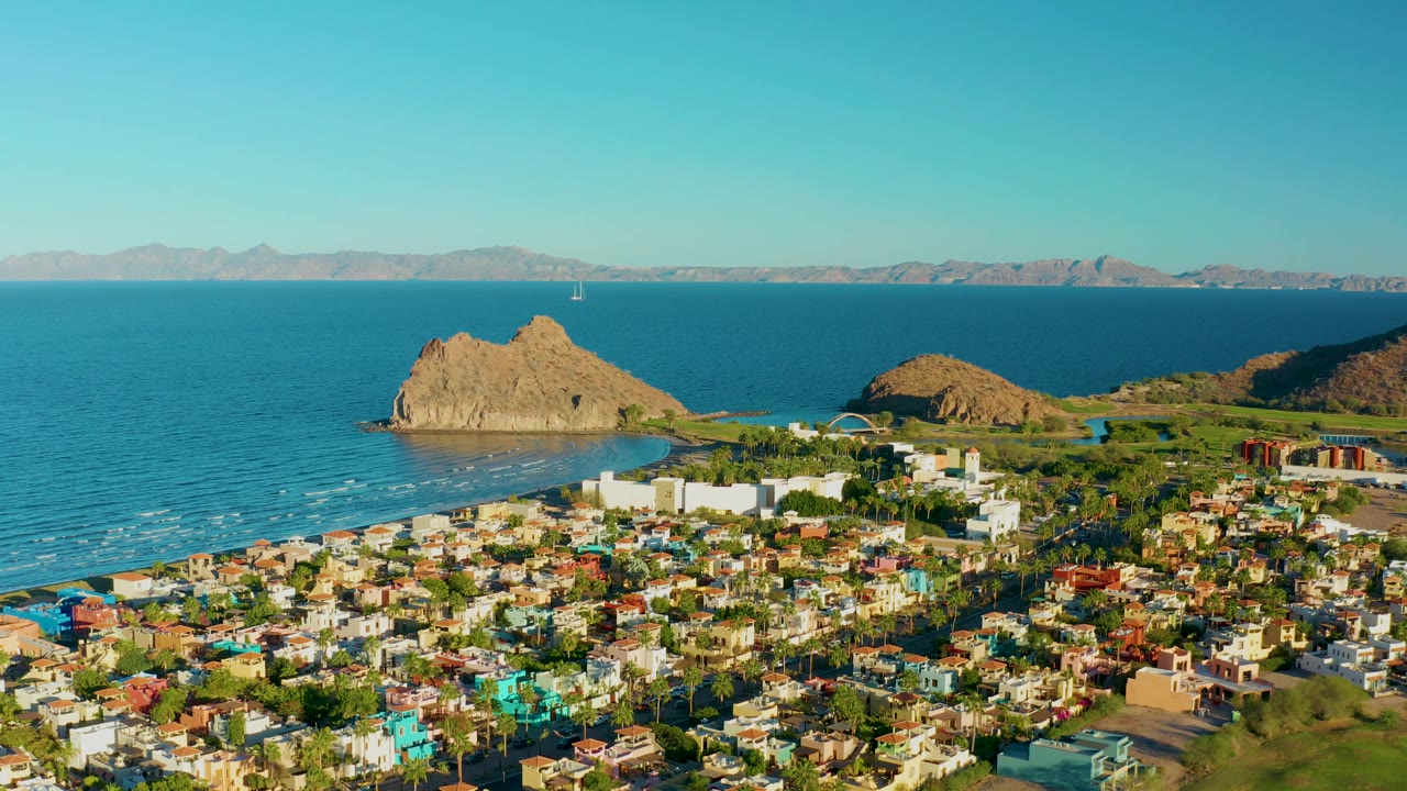 Aerial view in Loreto with the town spread across the lower frame, the rocky outcrop near the left-center coast, blue sea behind it, arid mountains on the horizon, and no visible people.