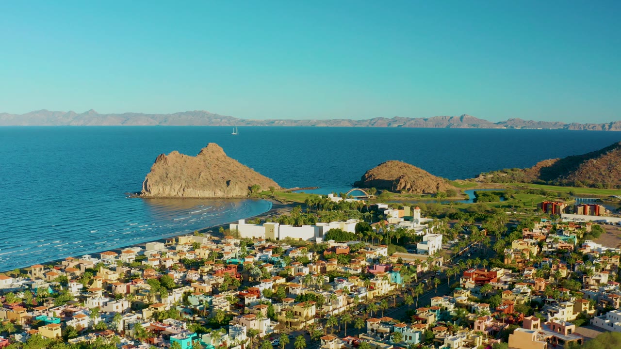 Aerial view over Loreto, Baja California Sur showing town rooftops, a white shorefront building complex, a curved beach, blue ocean, a rocky outcrop, distant mountains, and no visible people.