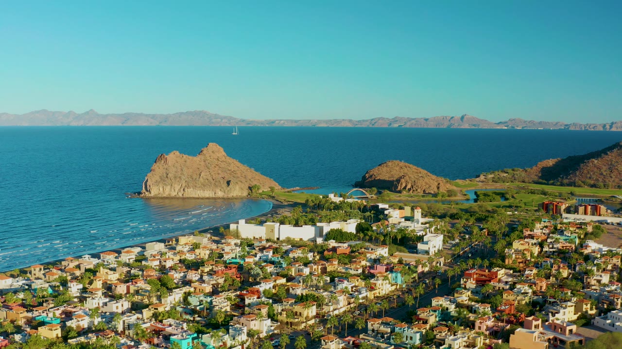 Aerial frame of Loreto with colorful rooftops in the lower foreground, white buildings near the beach, a tan rock formation offshore, blue water, mountain horizon, and no visible people.