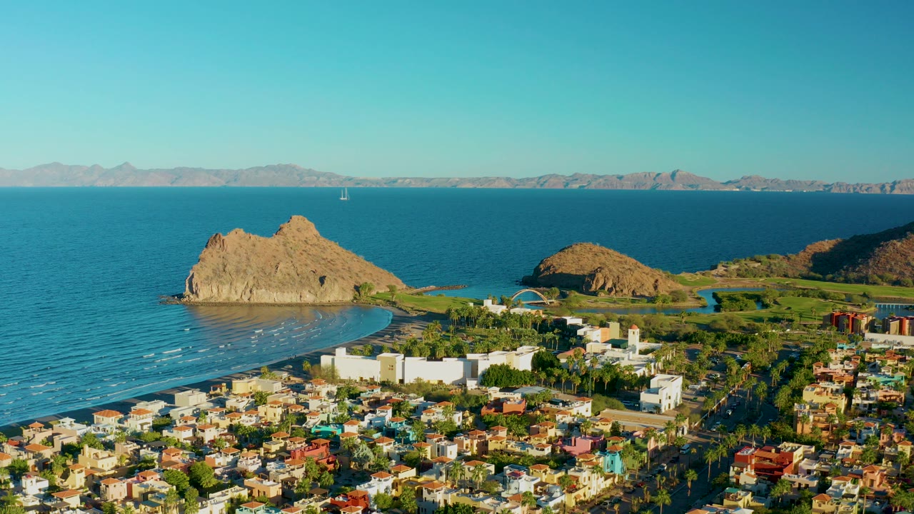 Opening aerial frame over Loreto, Baja California Sur, Mexico, showing town blocks in front of a blue bay, a large tan offshore rock, white resort buildings, distant mountains, and no visible people.