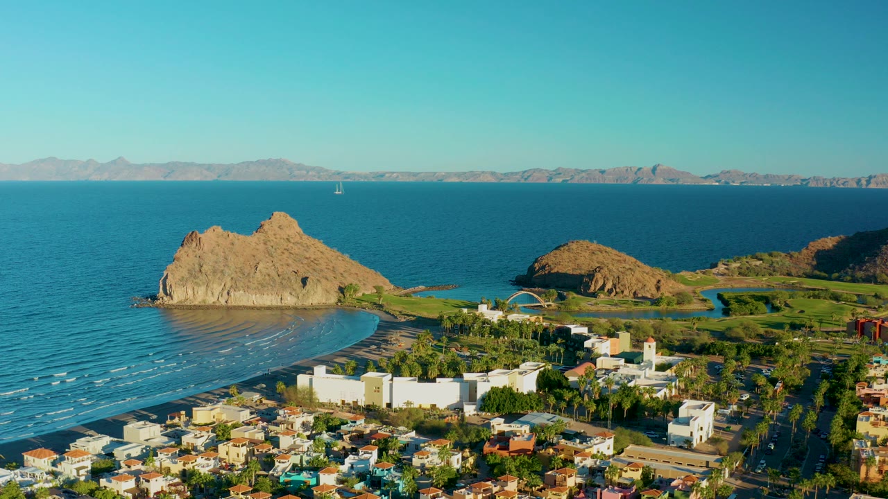 Aerial view over Loreto, Baja California Sur, with white town buildings in the foreground, a curved beach and tan rocky outcrop in the midground, blue ocean and distant mountains beyond, and no visible people.