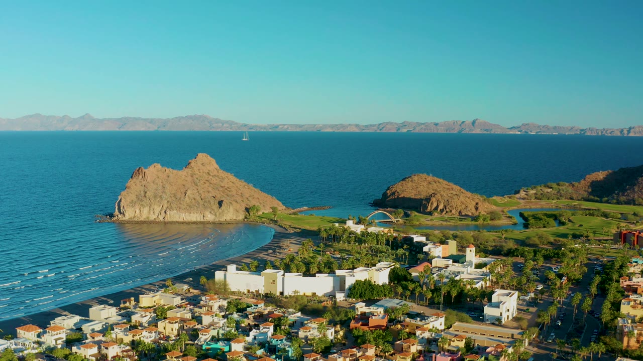 Coastal aerial with rocky outcrop, calm water, town buildings, and distant mountains.