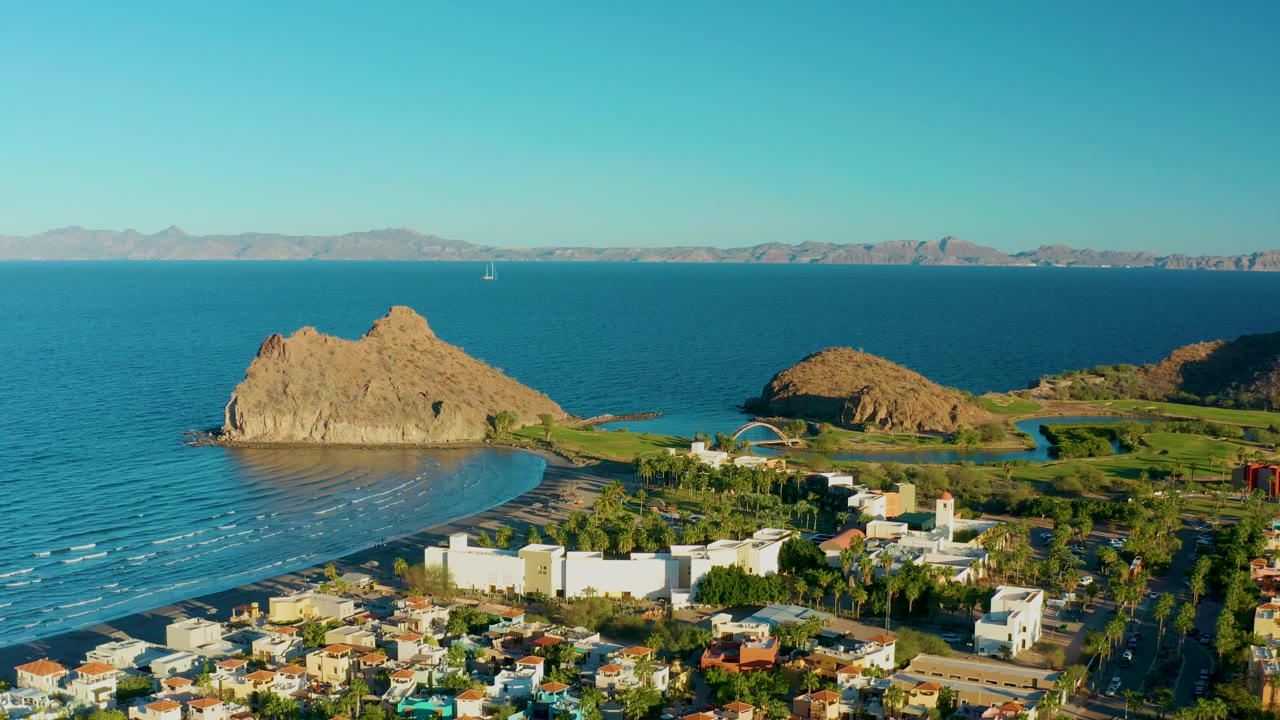 Drone view over Loreto with the beach and small wave lines at left, a large rocky outcrop near center, white town structures below, blue sea behind, and no visible people.