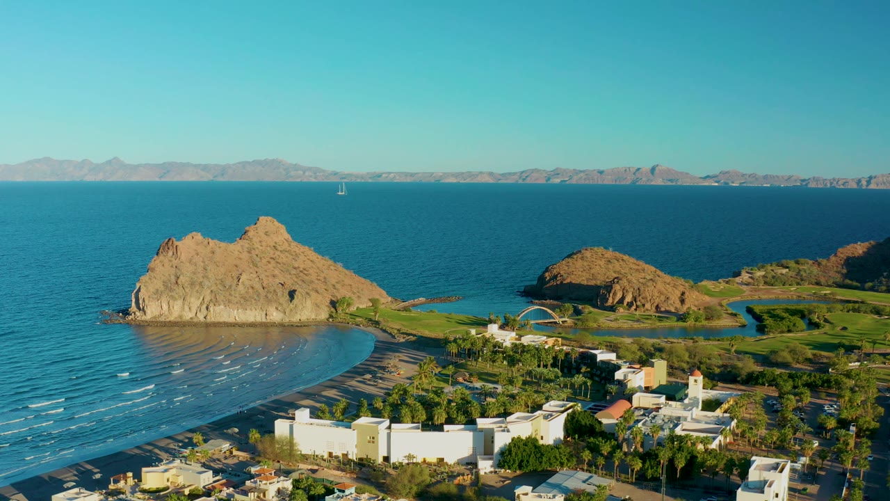 Early aerial frame over Loreto, Baja California Sur with small wave lines on the beach, the rocky outcrop left of center, coastal greenery and buildings below, and no visible people.
