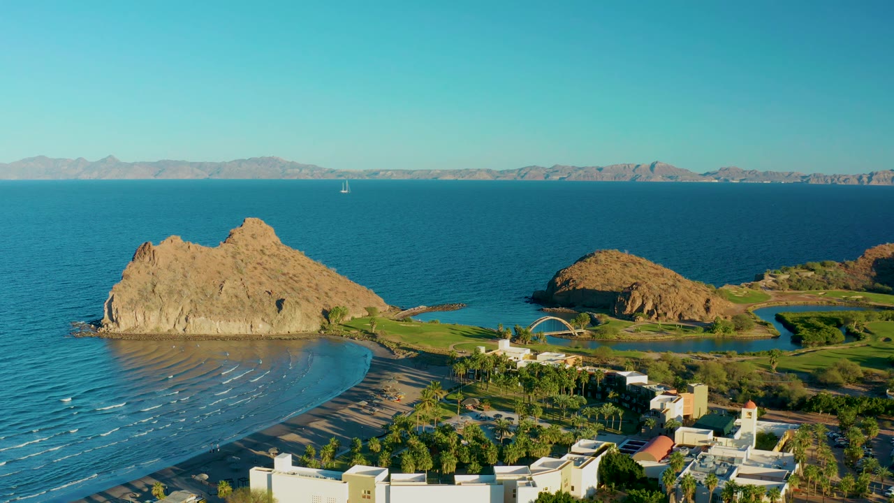 Aerial view in Loreto, Baja California Sur, showing small wave lines moving toward the sandy beach, a large tan rock outcrop, low buildings, palms, blue water, mountains, and no visible people.