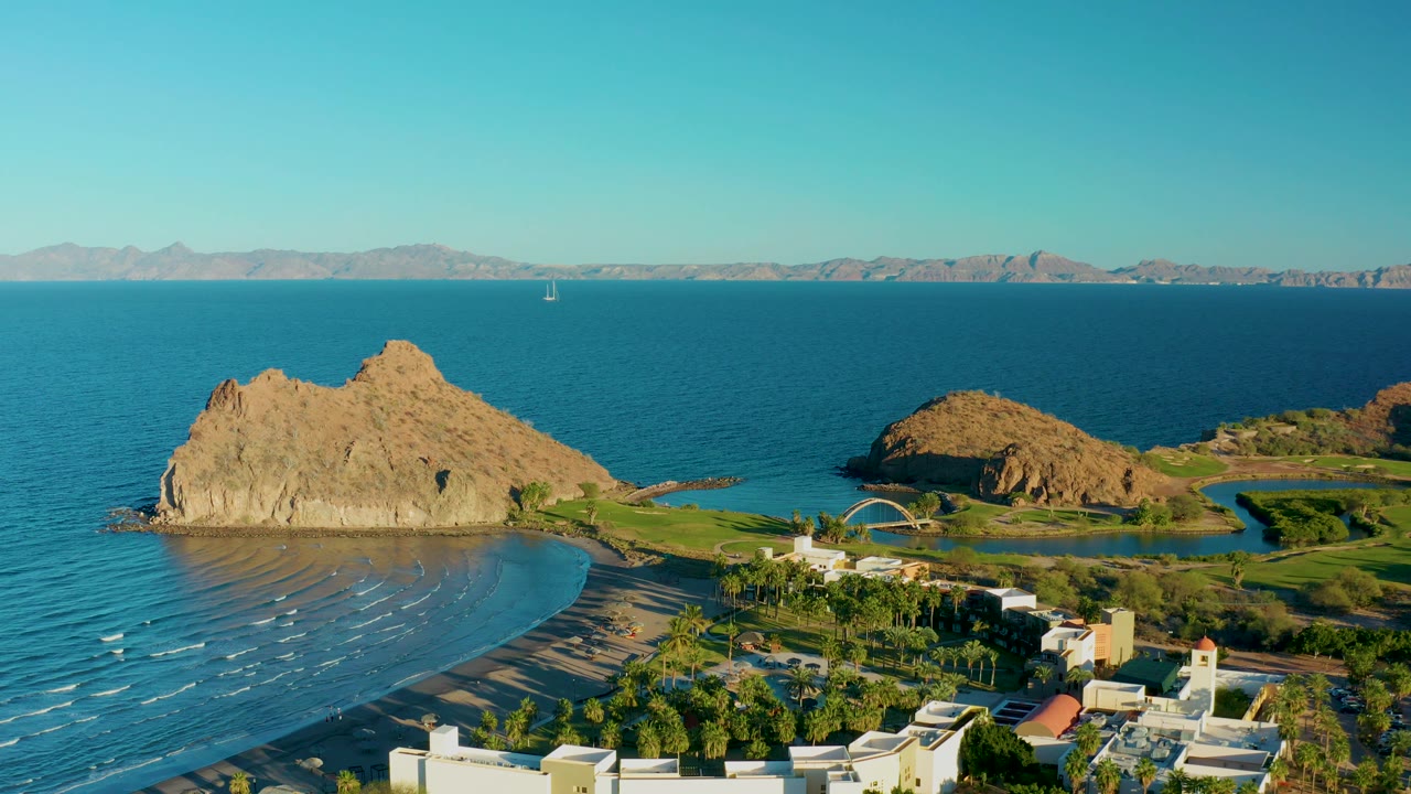 Mid-clip aerial frame in Loreto, Baja California Sur, looking across blue water and a tan rock formation toward a green shoreline area, low buildings, distant mountains, and no visible people.