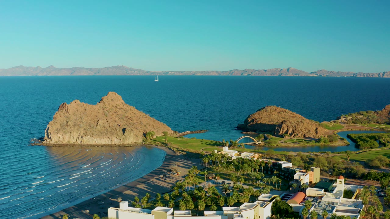 Aerial view in Loreto, Baja California Sur, Mexico, showing a tan rocky outcrop, calm blue ocean, curving beach, white beachfront buildings, distant mountains, and no visible people.