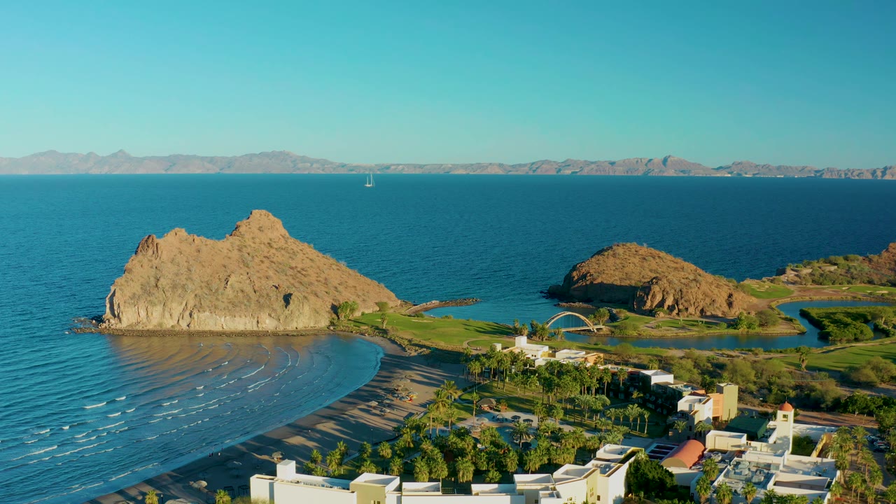 Aerial frame in Loreto, Baja California Sur showing the rocky outcrop slightly left of center, a crescent of wet sand and waves below it, foreground buildings and trees, open blue water, and no visible people.