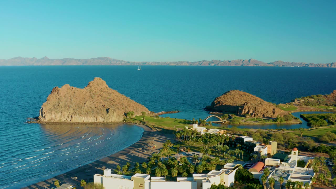 Mid-clip aerial view over Loreto, Baja California Sur with the rocky point, sandy beach, green shoreline areas, white coastal structures, broad ocean horizon, distant mountains, and no visible people.